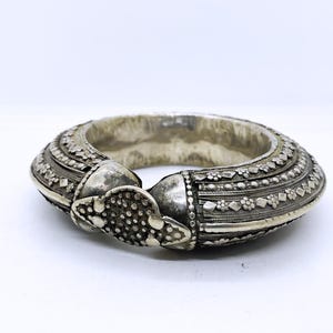 Può includere: Un bracciale a polsino argentato e ornato. Il bracciale presenta motivi dettagliati, tra cui disegni floreali e geometrici. La chiusura ha un elemento decorativo strutturato. Il gioiello è posto su uno sfondo bianco.