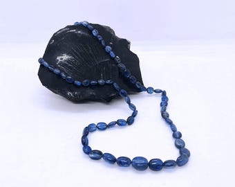 May include: A necklace of oval, deep blue beads rests on a dark, textured stone against a white background. The beads are strung together, forming a long strand. The necklace is a piece of jewelry.