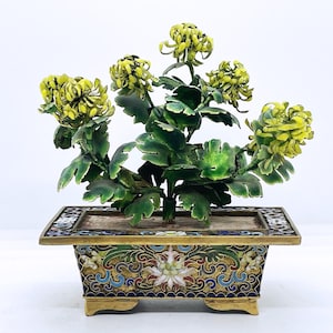 Peut inclure: Une composition florale décorative en cloisonné. La pièce présente des fleurs de chrysanthèmes jaunes et des feuilles vertes émergeant d'un pot rectangulaire orné. Le pot est décoré de motifs en émail coloré et d'accents dorés.