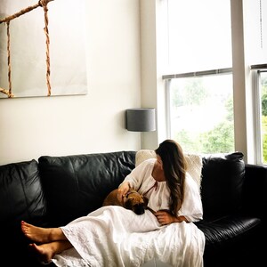 May include: A woman in a white dress sits on a black leather couch with a small brown dog. She is petting the dog and looking down at it. The couch is in front of a large window with a view of trees and a cloudy sky.