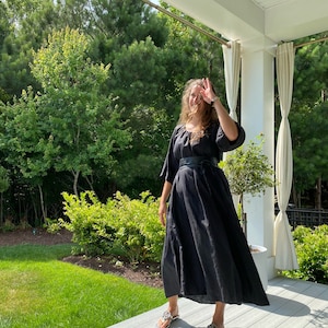 May include: A woman wearing a black maxi dress with a black belt and silver sandals. She is standing on a wooden porch with a green lawn and trees in the background.