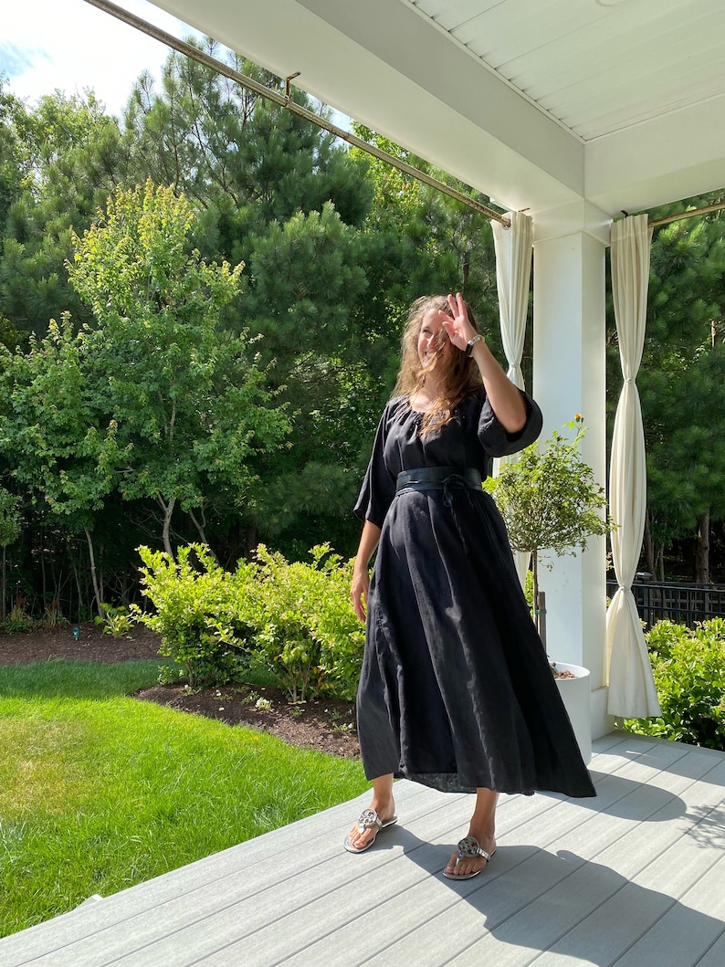 May include: A woman wearing a black maxi dress with a black belt and silver sandals. She is standing on a wooden porch with a green lawn and trees in the background.
