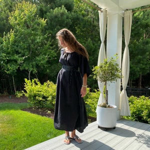 May include: A woman wearing a black maxi dress with a leather belt and sandals. She is standing on a wooden deck with a potted plant behind her.