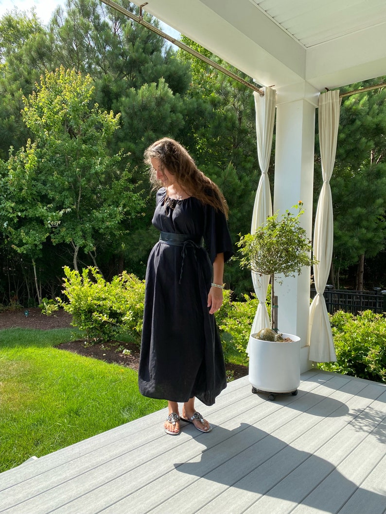 May include: A woman wearing a black maxi dress with a leather belt and sandals. She is standing on a wooden deck with a potted plant behind her.