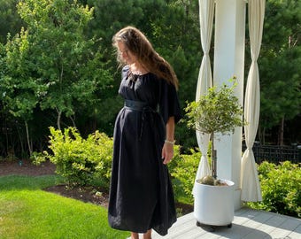 May include: A woman wearing a black maxi dress with a black leather belt and silver sandals. She is standing on a wooden deck with a potted plant behind her.