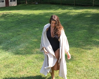 May include: A woman wearing a black swimsuit and a light beige linen robe walks on a grassy lawn.