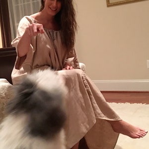 May include: A woman in a long, light brown dress sits on a bed with a white dog in front of her. The woman is smiling and looking at the dog.