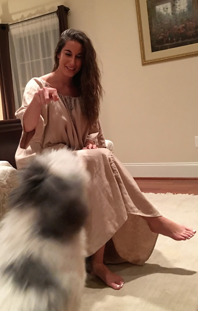 May include: A woman in a long, light brown dress sits on a bed with a white dog in front of her. The woman is smiling and looking at the dog.