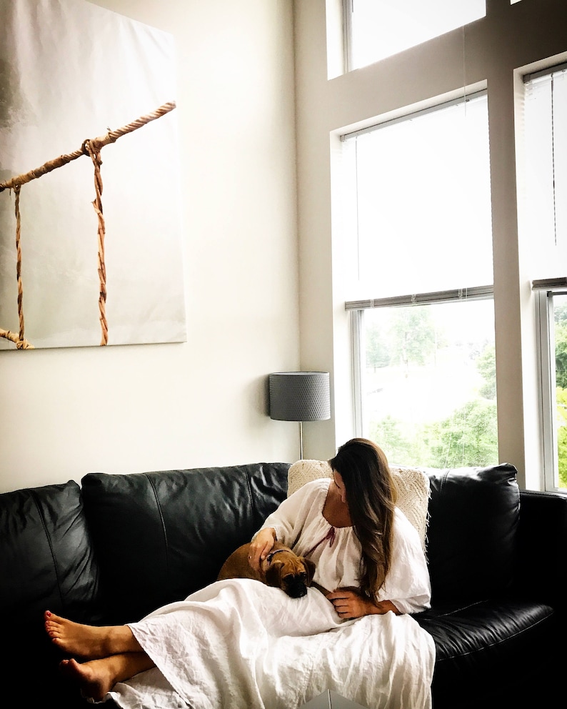 May include: A woman in a white dress sits on a black leather couch with a small brown dog. She is petting the dog and looking down at it. The couch is in front of a large window with a view of trees and a cloudy sky.
