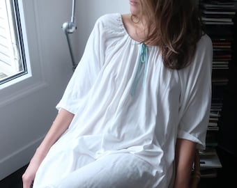 May include: A white, loose-fitting, long-sleeved dress with a gathered neckline and a light blue ribbon tie. The dress is worn by a person sitting on a chair in front of a window.