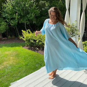 May include: A woman wearing a light blue, flowing maxi dress with a scoop neck and 3/4 sleeves. She is standing on a wooden deck with a grassy area and trees in the background.