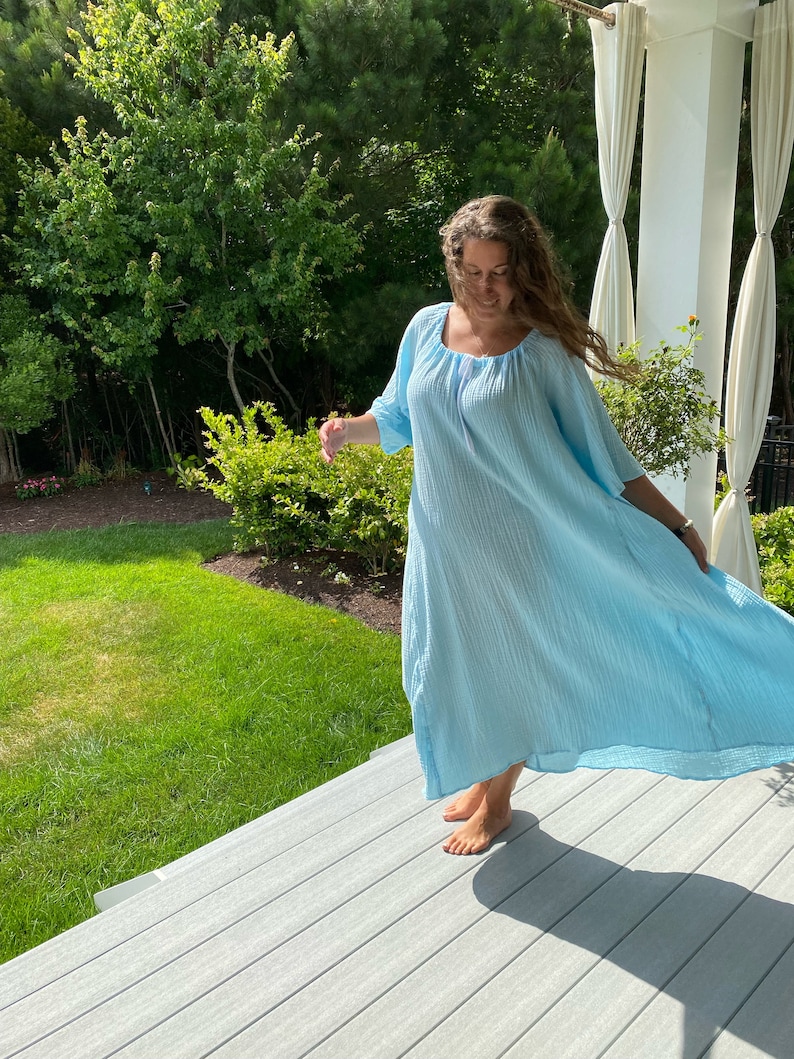 May include: A woman wearing a light blue, flowing maxi dress with a scoop neck and 3/4 sleeves. She is standing on a wooden deck with a grassy area and trees in the background.
