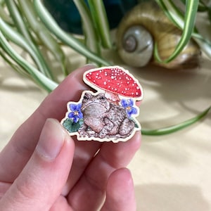 May include: A wooden pin featuring a toad sitting under a red and white mushroom with blue flowers. The toad is brown with a textured skin and has a surprised expression.