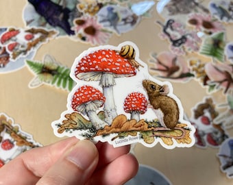 May include: A sticker featuring a whimsical illustration of a mouse, mushrooms, and a snail. The mushrooms have red caps with white spots. The mouse is brown and the snail has a yellow and brown shell. The sticker is signed "Lucinda Rowe".