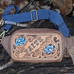 Puede incluir: Una riñonera de cuero marrón con una correa azul y un diseño de cuero repujado con el nombre "Faith" y flores azules.