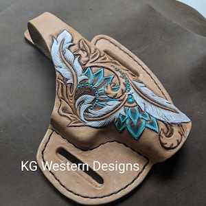 May include: A tan leather holster with a floral design featuring white feathers and turquoise accents. The holster is labeled "KG Western Designs".