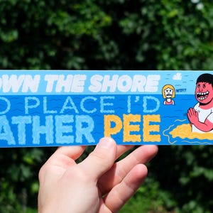 May include: A rectangular sticker with a blue background and white text that reads "DOWN THE SHORE NO PLACE I'D RATHER PEE." The sticker features cartoon illustrations of people in the water. The sticker is held by a hand.