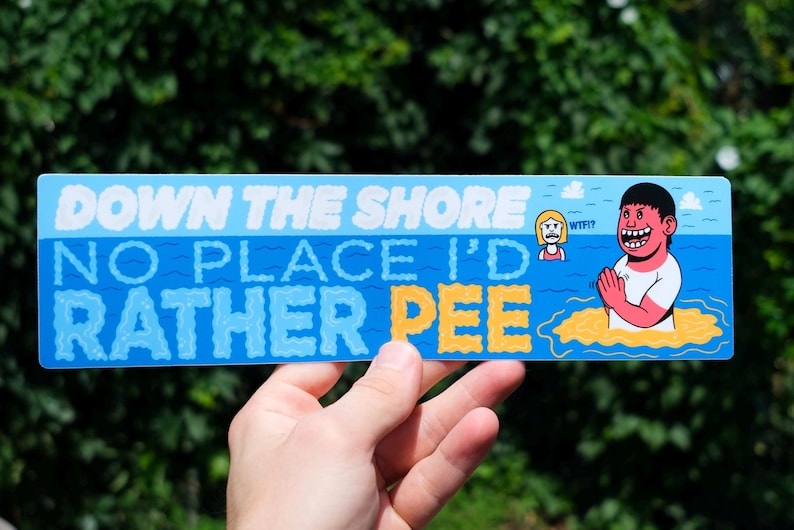 May include: A rectangular sticker with a blue background and white text that reads "DOWN THE SHORE NO PLACE I'D RATHER PEE." The sticker features cartoon illustrations of people in the water. The sticker is held by a hand.