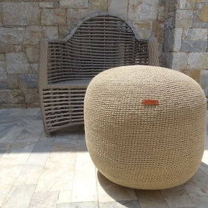Op de afbeelding: Een grote, ronde, beige gebreide pouf ottomane staat op een stenen terras naast een rieten stoel.
