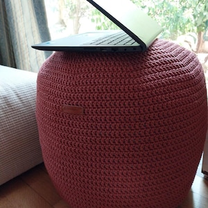 Könnte beinhalten: Ein großer, runder, rot-oranger gehäkelter Ottoman mit einem Laptop darauf. Der Ottoman ist aus dickem Garn gefertigt und hat eine strukturierte Oberfläche. Der Laptop ist schwarz und geöffnet und zeigt die Tastatur. Der Ottoman steht auf einem Holzboden.