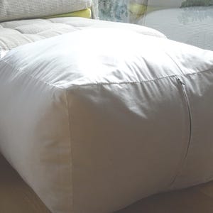 May include: A square, off-white ottoman with a zip detail. The ottoman is made of a soft fabric and is placed in front of a light-coloured sofa. The design is simple and modern, suitable for various interior styles.
