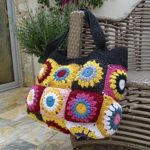 Può includere: Una borsa tote in crochet nero con toppe colorate a quadrati della nonna. La borsa ha un'unica tracolla ed è appoggiata su una sedia in vimini.