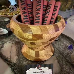 Puede incluir: Un cuenco de madera lleno de caramelos de regaliz con forma de dinamita roja y negra. Los caramelos están envueltos en papel rojo con letras negras que dicen "Dynamite". Un cartel debajo del cuenco dice "Dynamite (regaliz)".