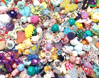 Puede incluir: Un colorido surtido de cuentas y abalorios de diversas formas y tamaños. La colección incluye cuentas con forma de corazón, flor y animales en tonos rosa, azul, amarillo y morado. Algunas cuentas tienen superficies brillantes o texturizadas, creando una mezcla vibrante.