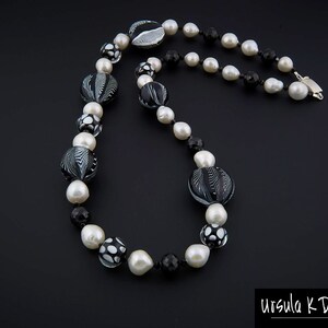 Pu&ograve; includere: Una collana di perle di vetro nere e bianche con una variet&agrave; di motivi, tra cui spirali, punti e forme geometriche. La collana &egrave; realizzata con perle grandi e piccole ed &egrave; infilata su una catena d'argento.