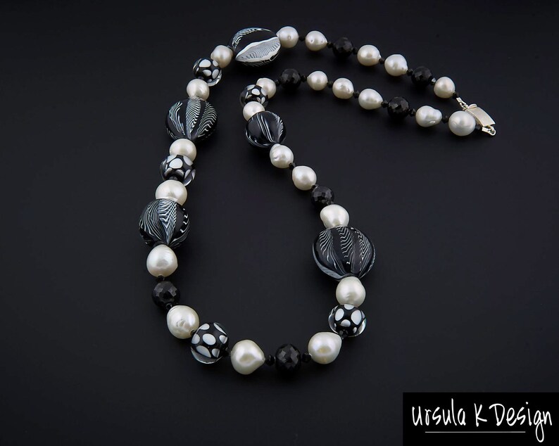 Pu&ograve; includere: Una collana di perle di vetro nere e bianche con una variet&agrave; di motivi, tra cui spirali, punti e forme geometriche. La collana &egrave; realizzata con perle grandi e piccole ed &egrave; infilata su una catena d'argento.