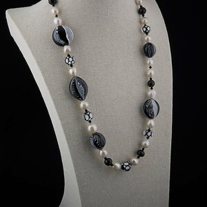Pu&ograve; includere: Una collana bianca e nera con grandi perline di vetro con motivi e perle bianche. La collana &egrave; esposta su un manichino bianco.