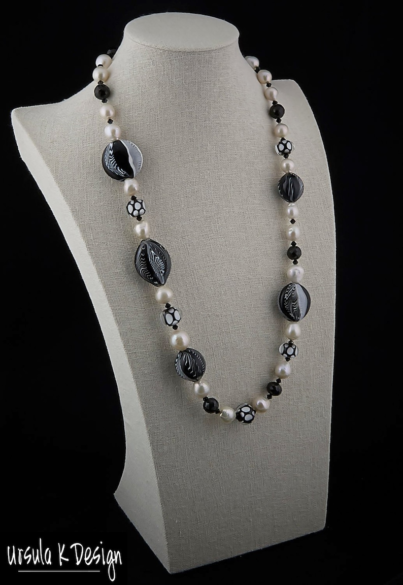 Pu&ograve; includere: Una collana bianca e nera con grandi perline di vetro con motivi e perle bianche. La collana &egrave; esposta su un manichino bianco.
