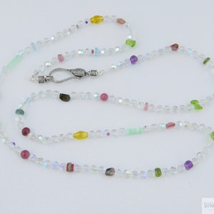 Może przedstawiać: Długi naszyjnik z kryształów z srebrnym zapięciem i kolorowymi koralikami z kamieni szlachetnych. Koraliki są w odcieniach różowego, zielonego, żółtego i fioletowego.