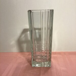 May include: A clear glass vase with a square shape and a geometric pattern. The vase is empty and sitting on a pink striped surface.