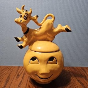 May include: A yellow ceramic cookie jar featuring a round base shaped like a smiling face with closed eyes and a curved mouth. A sculpted yellow cow with black hooves and a black collar is positioned on top of the lid, appearing to jump.