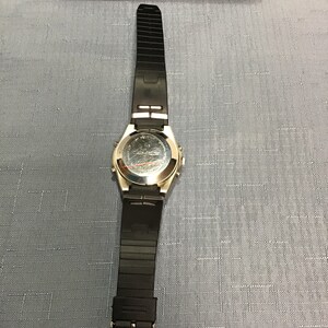 May include: A silver-toned wristwatch with a black rubber strap. The watch has a round face and a silver-toned bezel. The watch is on a light blue surface.