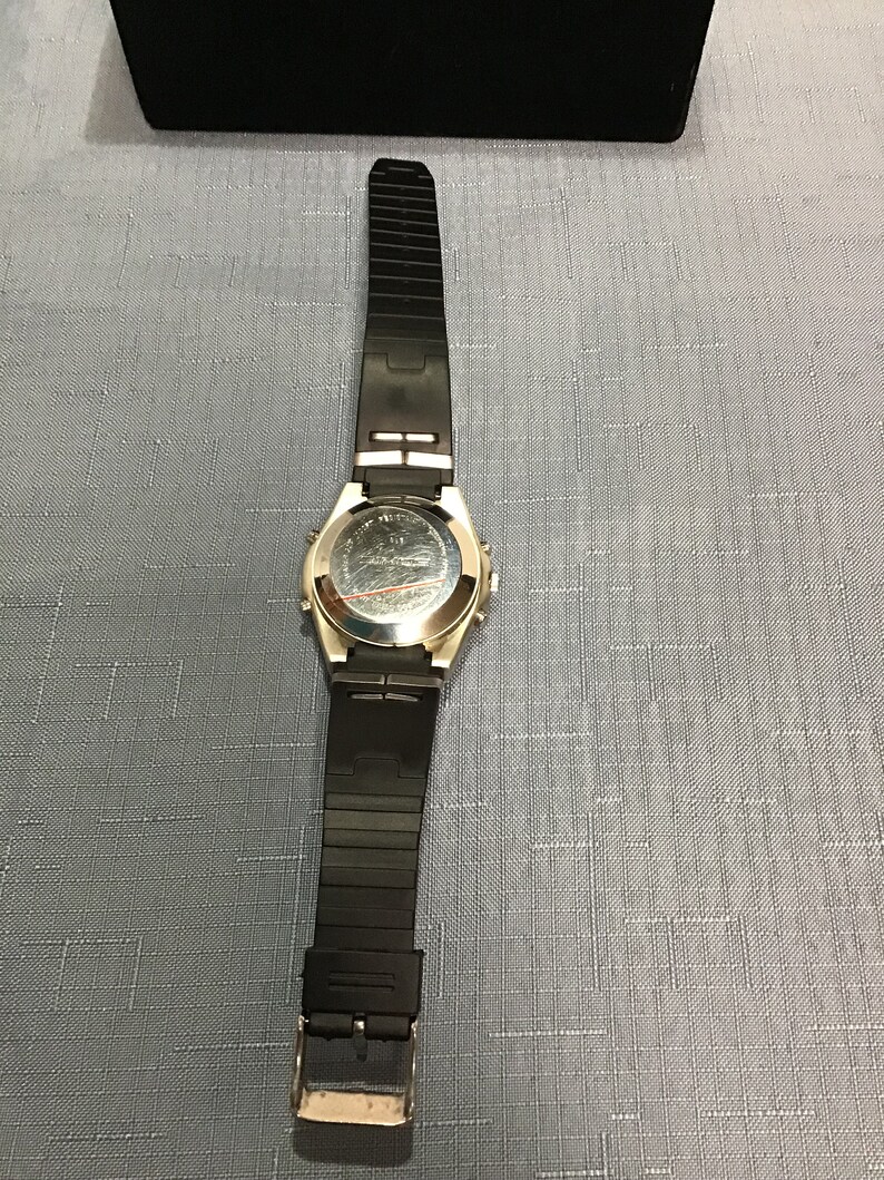 May include: A silver-toned wristwatch with a black rubber strap. The watch has a round face and a silver-toned bezel. The watch is on a light blue surface.