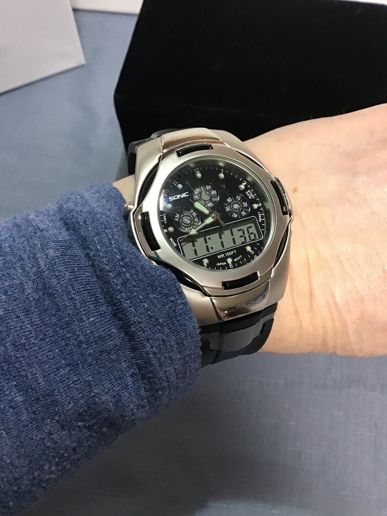 May include: A silver and black digital wristwatch with a black face and a silver band. The watch has a black digital display that reads "11:13:36" and the words "WR 100FT" and "SONIC" are visible on the face.