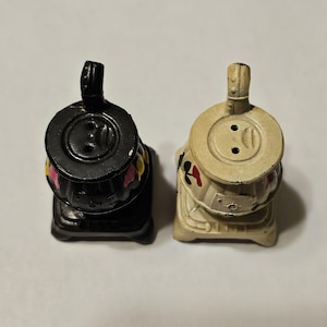 May include: Two miniature stove-shaped salt and pepper shakers. One is black, the other is off-white. Both have a chimney and decorative floral accents in pink, yellow, and red. The shakers are on a plain background.