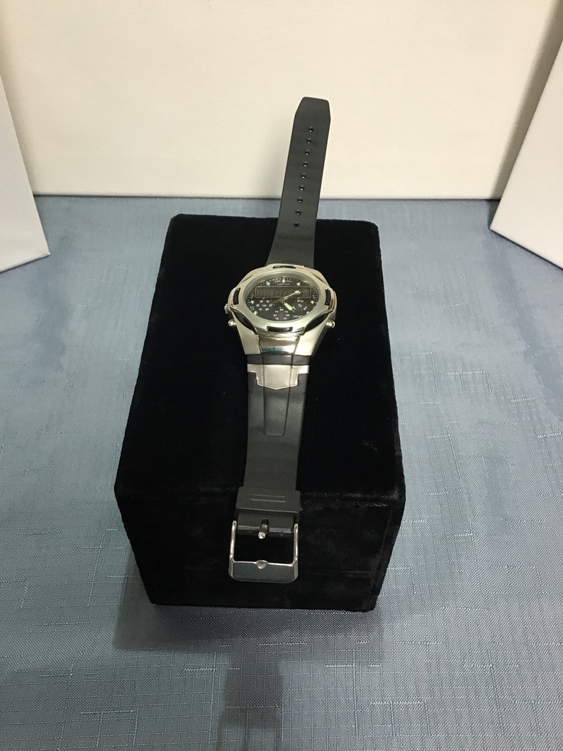 May include: A silver and black wristwatch with a black rubber strap. The watch has a digital display and is sitting on a black velvet box.