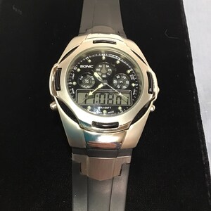 May include: A silver and black digital wristwatch with a black rubber strap. The watch face has a black background with white numbers and a silver bezel. The watch face also has a small analogue clock with a black face and silver hands. The watch is water resistant to 100 feet. The watch face displays the time, date, and day of the week.