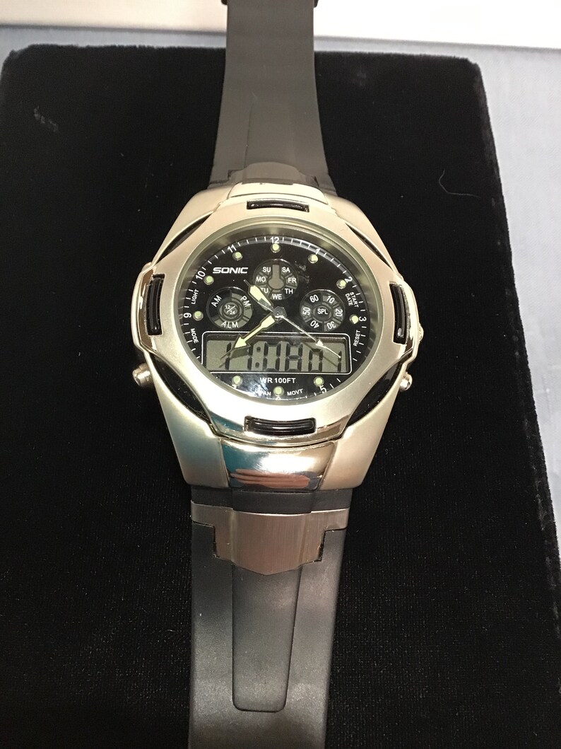 May include: A silver and black digital wristwatch with a black rubber strap. The watch face has a black background with white numbers and a silver bezel. The watch face also has a small analogue clock with a black face and silver hands. The watch is water resistant to 100 feet. The watch face displays the time, date, and day of the week.