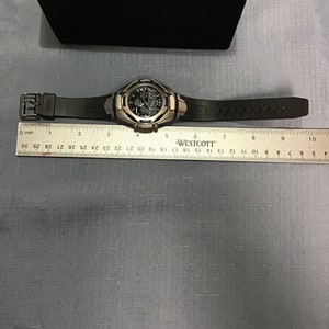 May include: A black digital watch with a silver metal band and a black rubber strap. The watch has a black face with white numbers and a silver metal bezel. The watch is on a ruler with the text "WESTCOTT" printed on it.