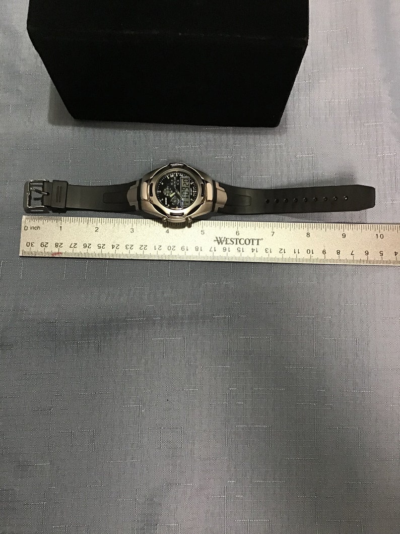 May include: A black digital watch with a silver metal band and a black rubber strap. The watch has a black face with white numbers and a silver metal bezel. The watch is on a ruler with the text "WESTCOTT" printed on it.