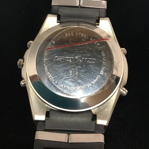 May include: Rear view of a silver Sonic watch with a black rubber strap. The watch face has the text "Sonic" and "Stainless Steel Back" printed on it. The watch is water resistant.