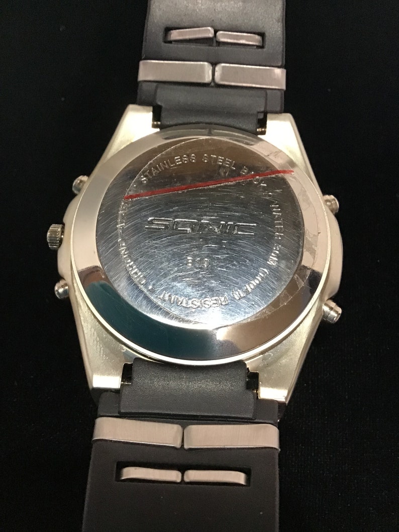 May include: Rear view of a silver Sonic watch with a black rubber strap. The watch face has the text "Sonic" and "Stainless Steel Back" printed on it. The watch is water resistant.