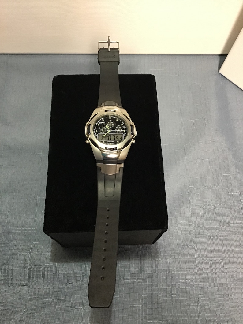 May include: A silver and black wristwatch with a black rubber strap. The watch face has a digital display and analogue hands.