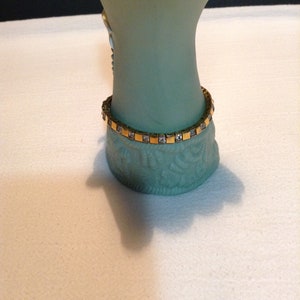 May include: A gold tone bracelet with clear square-shaped stones. The bracelet is on a light green surface.