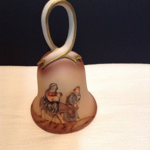 May include: A ceramic bell with a white and gold handle. The bell is decorated with a scene of the nativity, featuring Mary, Joseph, and the baby Jesus riding on a donkey.