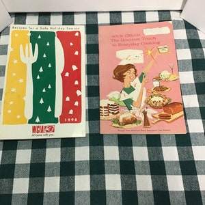 May include: Two vintage cookbooks with colorful covers. The cookbook on the left has a red, yellow, and green cover with a fork, knife, and spoon graphic. The cookbook on the right has a pink cover with a woman holding a whisk and a bowl of sour cream. The text on the cover reads "Sour Cream: The Gourmet Touch to Everyday Cooking".